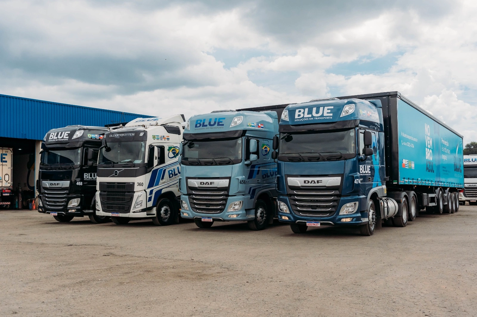 Quatro cavalos da BLUE alinhados no pátio, diante do galpão azul: DAF preto 468, Volvo branco 360, DAF azul-claro 456 e DAF azul-marinho 474 com a carreta do Novembro Azul.
