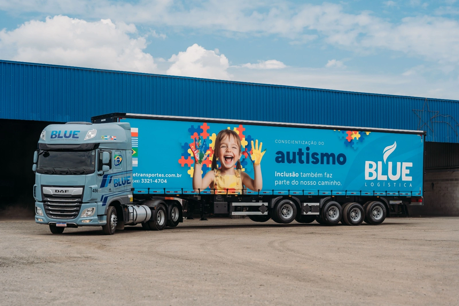 Caminhão DAF azul-claro da BLUE com carreta lonada azul da campanha de conscientização do autismo, com a foto de uma criança sorrindo entre peças de quebra-cabeça.