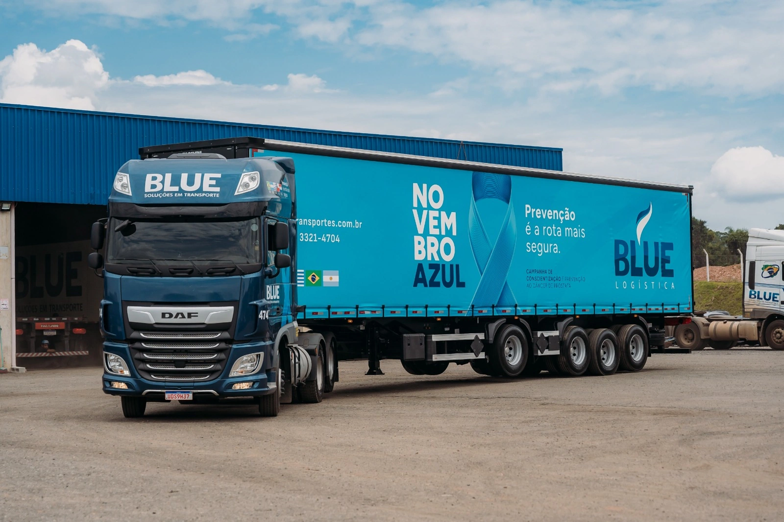 Caminhão DAF azul-marinho da BLUE com carreta lonada azul da campanha Novembro Azul, saindo do galpão.